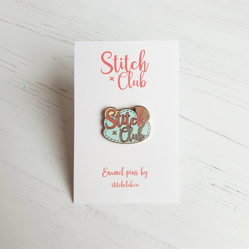 A hard enamel pin badge attached to a card backing. It features the logo of Stitch Club sewing kit designers.