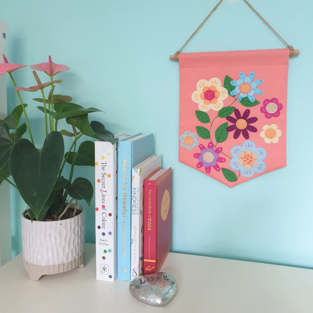A colourful handsewn applique banner, decorated with felt flowers, hangs on a turquoise wall. On a surface in front, sits a plant and some books.