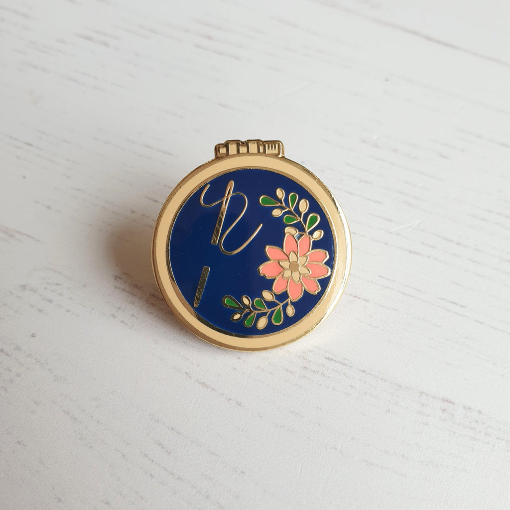 A hard enamel pin badge featuring an embroidery hoop design with navy background and floral details with a gold sewing needle.