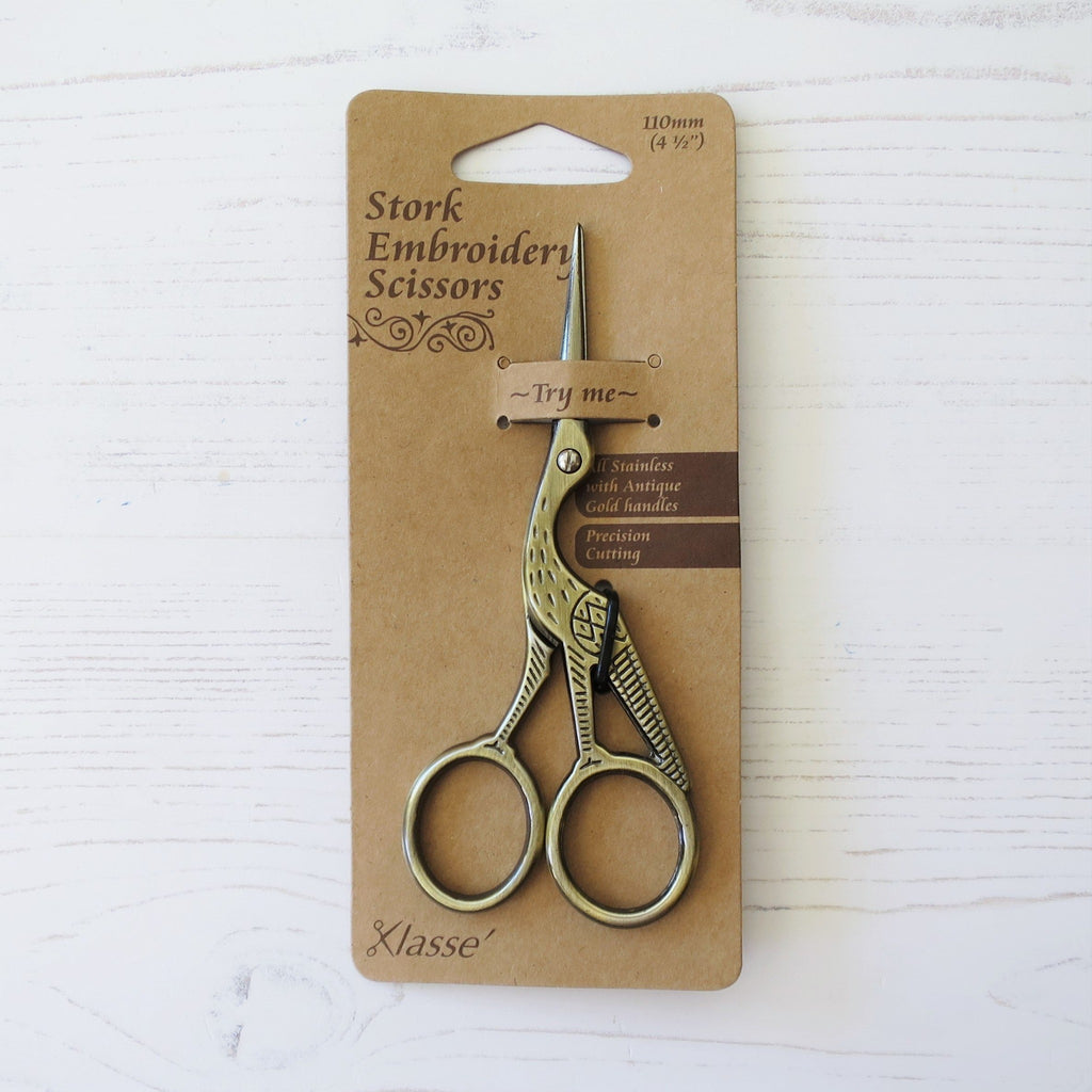 A pair of stork embroidery scissors, brass coloured and with a kraft cardboard backing.