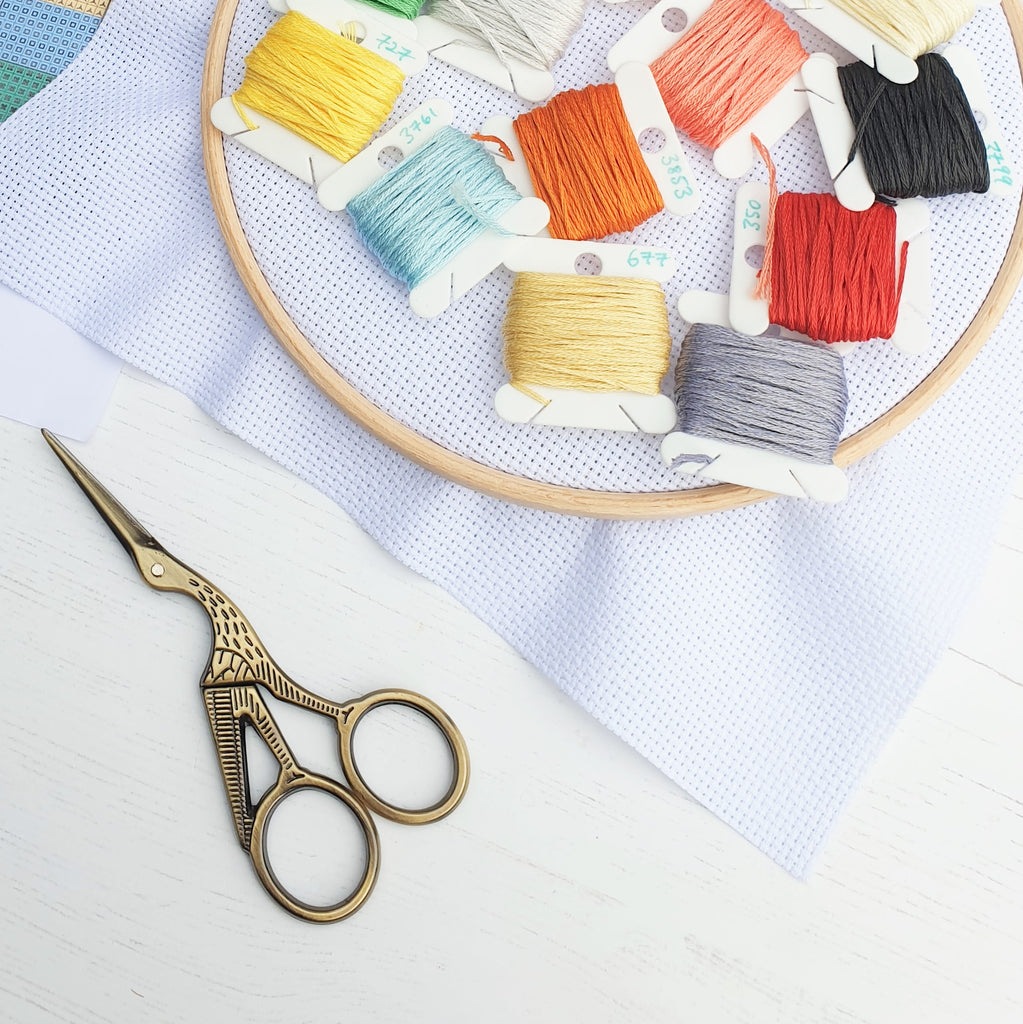 Colourful sewing bobbins wound with brightly coloured embroidery threads, resting on cross stitch fabric in a hoop, and also with a pair of stork sewing scissors.