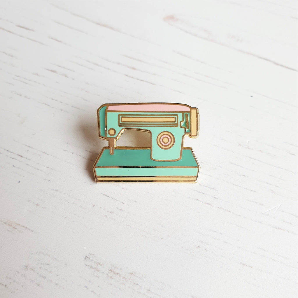 A hard enamel sewing machine pin badge, featuring and 1950s style sewing machine in mint greens and pink with gold plating.